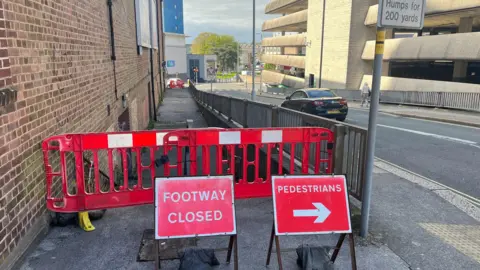 Two red signs with the words 'footway closed' and 'pedestrians' in front of a red barrier around the drain.