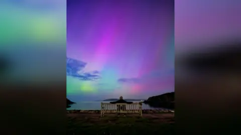 Mathew Browne A man is sat on a bench with his arms sprawled out either side as he looks up at the Northern Lights illuminating the night sky in purple, blue and green colours. In front of him is the west Wales coast.