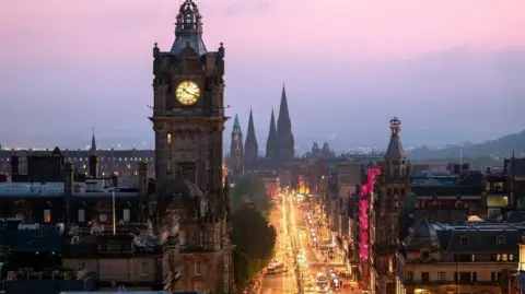 Joe Daniel Price/Getty Images The Balmoral Hotel clock rises into a purple night sky next to a lit-up  Princes Street, glowing golden from the street lights. We can see church spires at the far end and other buildings line the other side of the street. The picture was taken from height so we are looking down on the street.