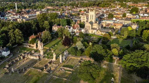 An aerial image of the sand-coloured St Edmundsbury Cathedral, which has a square tower with tall spikes at each corner. In the foreground is are grey stumps which are the ruined masonry of a medieval abbey. The site is surrounded by trees and in the background the buildings of a town centre. 