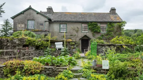 National Trust Hill Top, a grey farmhouse with green windows. There's a garden in front of the house with various green plants.