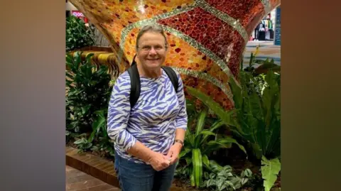 Supplied A woman wearing a patterned top with a rucksack on her back standing in front of a colourful and patterned mosaic-style structure amongst some long green leaves. She is smiling. 