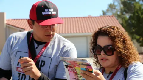 Olga Mexia y otro miembro de la Unión Culinaria recorriendo un vecindario en el norte de Las Vegas a finales de septiembre. Olga lleva gafas de sol y lee el texto de un folleto.