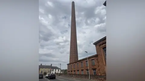 Dixon's Chimney. It is a red brick structure that towers above all other buildings in the area. Several cars are stopped at nearby traffic lights. The sky is filled with clouds.