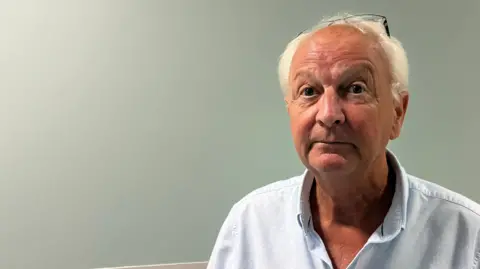 Robert Wright with a pale blue shirt in the corridor of the new emergency care unit at Arrowe Park Hospital. He has white hair with his glasses resting on his head. He looks impassively at the camera.