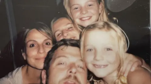 Chris Strong A family photo of the Bastion-Strong family showing Chris and Katie with their three daughters. The family are all close together and smiling as they pose for the picture.