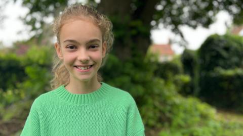 Anna, an 11 year old girl, wears a green jumper and stands in front of woodland