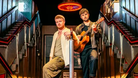 Tommy Ga-Ken Wan Two men, dressed casually, on a stage set showing twin flights of stairs. The man on the right has an acoustic guitar.
