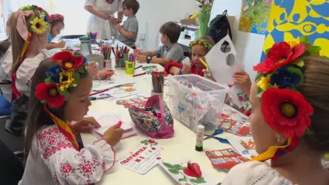 Several children from Ukraine wear traditional folk clothing from their homeland while doing work at a Saturday school lesson. The children, some of whom are wearing flowers in their hair, are colouring in pieces of paper.