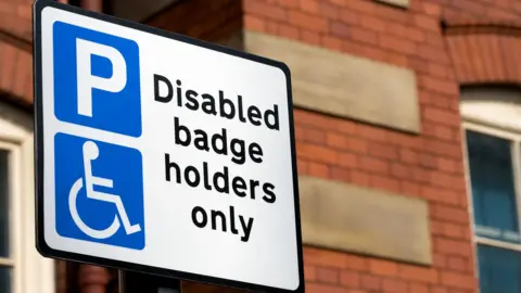 A road sign showing parking is only for disabled badge holders. Behind is a red-brick house. 