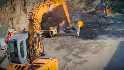 Two people, whose identities have been blurred, sit in a the bucket of a yellow bucket on the site.