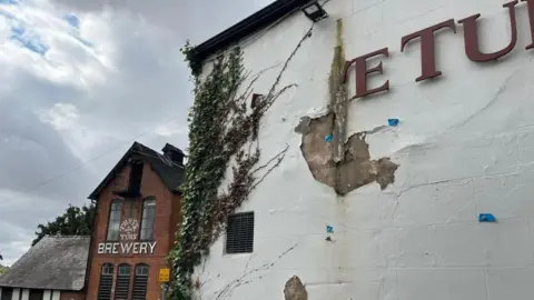 In the foreground is the front wall of the Three Tuns Inn - the white plaster is cracked and peeling off, and the sign is falling apart, so it just reads 'E TUN' in dark red lettering. Ivy is growing up part of the wall. In the background, the Three Tuns Brewery can be seen with its name painted on the red brick building in white lettering.