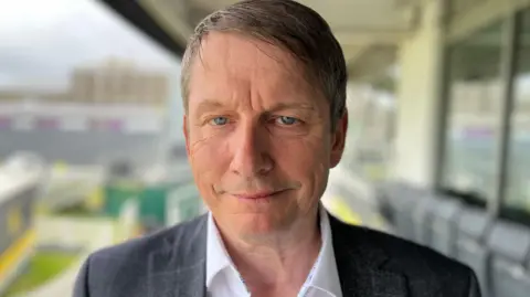 Peter Matthews. He has short grey hair and blue eyes. He is wearing a white shirt and a grey suit jacket. He is looking at the camera and smiling.