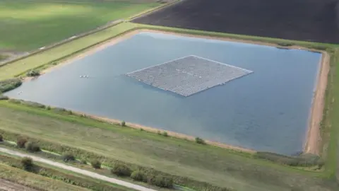 A large square reservoir of water in the middle of a field with a road in the foreground. 