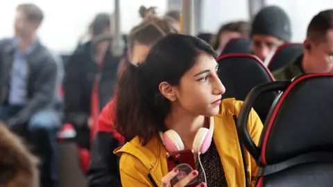 A young woman wearing a yellow jacket and pink headphones is holding a mobile phone while sitting on a bus.