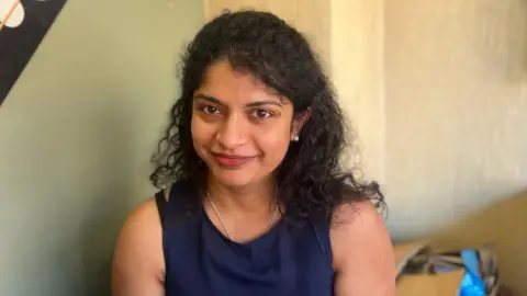 A mid shot of a woman who has brown, curly hair and is wearing a navy blue vest top. She is smiling into the camera and is sitting in front of a cream wall. 