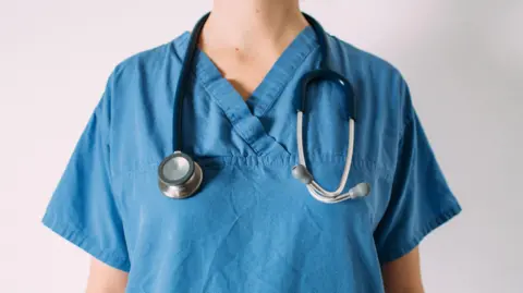 A woman in blue medical scrubs. There is a stethoscope hanging around her neck. 