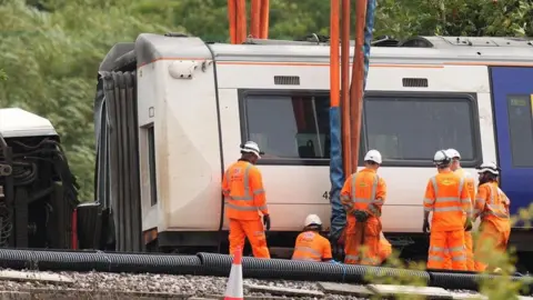 Eddie Mitchell Recovery workers in orange high-visibility clothing and white safety helmets stand beside a Southern train carriage as lifting equipment is attached. Thick straps hang from a crane above the carriage, while cables, pipes and railway infrastructure are visible in the foreground.