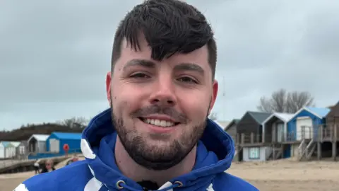 Ruben smiling at the camera, wearing a blue hoodie in front of a row of beach houses