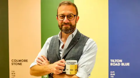 A bearded man with glasses holding a white mug with yellow stripes. He is wearing a shirt and tweed waistcoat. Behind him are colour strips of stone, green, yellow and blue.