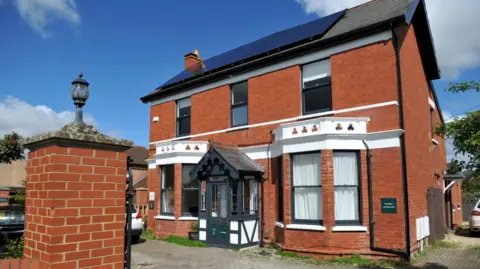 Angie Petkovic A large Victorian or Edwardian style detached suburban house on a sunny day. There are large bay windows on the bottom storey and a Tudor style porch. There are clouds up above. Some cars can be seen parked on the driveway.