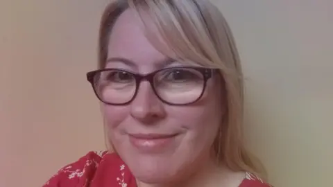 Nicola Armstrong A woman smiling at the camera, she has blonde hair, glasses and is wearing a red and white top