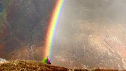 a hillwalker sits at the top of a peak with a rainbow appearing to come down on their head