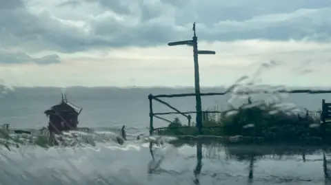 Sarah n Essie View of a heavy shower of rain seen through a window. The glass and the water slightly distort the seaside scene featuring signpost, a set of metal railings and a small shed-like building with a pitched slate roof.