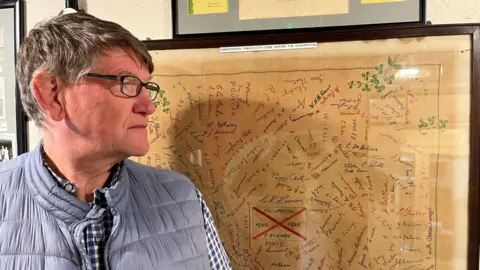 Damien Horn looking at a table cloth covered with embroidered signatures. The tablecloth is worn and behind a frame.