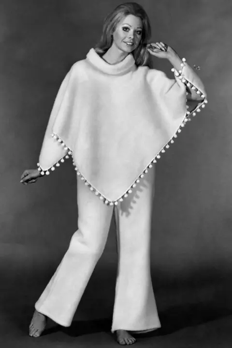 Getty Images Black and white advertising photo of a woman wearing "poncho pyjamas" - a white poncho trimmed with bobbles and flared trousers. The model has bare feet, heavy eye makeup, and one arm aloft. 