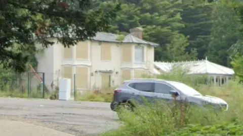 The Woodlands Hotel is seen off centre on the left, with two bay windows on each side. The building, which is an off white colour, has its windows and doors boarded up. A silver car is parked in the car park just outside of the hotel. 