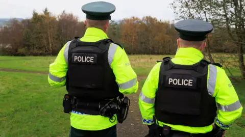 PA Media Two police officers stand in a park with their backs to the camera. They are wearing bright yellow coats and a black police branded vest. The park they are standing in has a lot of trees and a winding path leading off to the left.