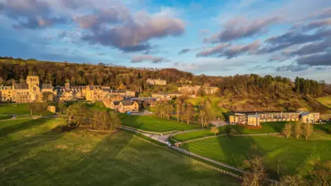 Getty Images View of buildings spread across grounds, with trees and playing fields. Behind the buildings is a wooded area. Blue skies and clouds.