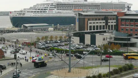 Graham Jones Image shows the mast standing on a roundabout near the pierhead in Liverpool. The Queen Mary 2 cruise ship is taking up much of the photograph's background as it berths on the Mersey. 