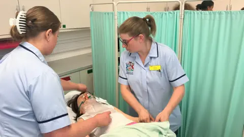 Two nursing students work around a female dummy, who is wearing an oxygen mask, behind a curtain in a university's simulated hospital wing during a practical