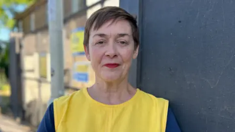 A woman with short dark hair looks at the camera. She is wearing a bright yellow tabard 