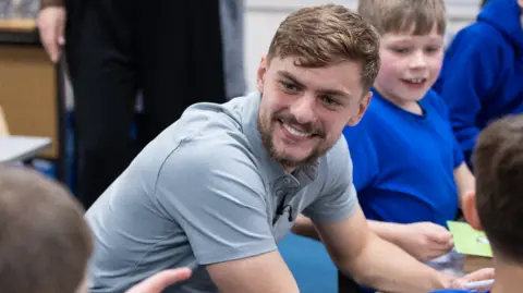 Everton Kiernan smiling in a grey polo shirt in the classroom.