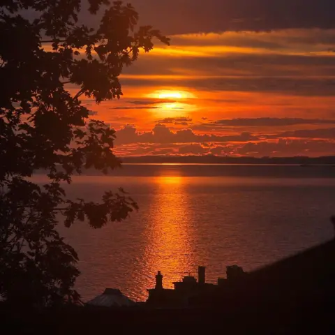 Fred Tordeur An orange sunrise with the bright beam reflected in the sea, rooftops and trees are silhouetted in the foreground.