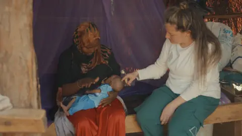 Jess Hallett (right) pictured with one of the refugees and their baby. She strokes the babies head while the mother breastfeeds the baby. 