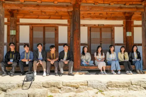 BBC/ Hosu Lee A group of five men and five women wearing lanyards sit along a wooden porch of a traditional Korean temple with large timber beams and lattice doors. The group is spread across the veranda in bright daylight, with a stone foundation in the foreground and a bag resting near the front edge.