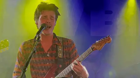 @jmericks | Instagram Cruz Beckham performing on stage at Clwb Ifor Bach. He is playing the guitar with his eyes shut and singing into a microphone. He hears a colourful patterned shift and has short dark hair.