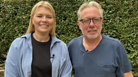 A smiling man and woman stood side by side. The woman has long blonde hair and is wearing a blue open shirt with a black t-shirt underneath. The man is wearing glasses and a blue t-shirt. They are standing in an outdoor setting with a green bush behind them. 