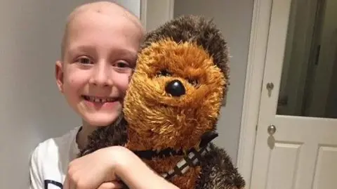 Ryan Quinn Aaron, his hair wispy and fluffy after treatment, smiles broadly as he hugs his well-loved Chewbacca soft toy. He wears a white t-shirt and is in a grey hallway with white doors.