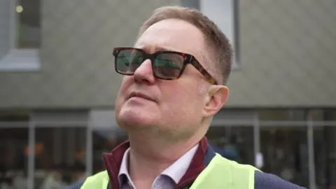 A man wearing sunglasses standing outside a large building. He has a florescent jacket over a blue jacket and a pink shirt. He has close cropped brown hair.