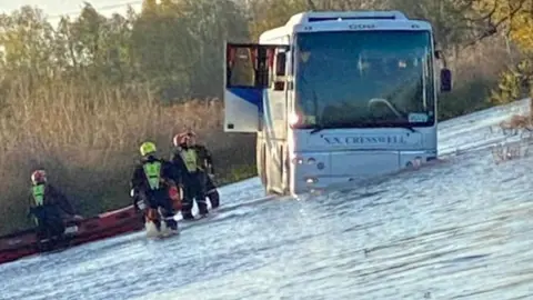 A bus stuck in flood water with a door open. On the left of the photo, near the bus, are at least three firefighters and a small boat. 