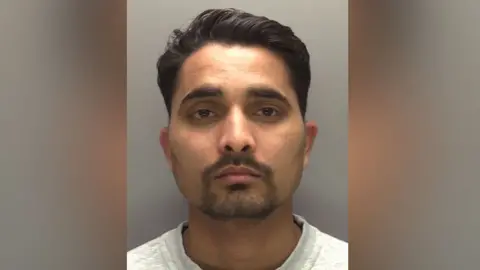 A head‑on police mugshot of Prajapati with short, dark, neatly styled hair and a trimmed beard and moustache. He has a neutral expression, medium skin tone, and is wearing a light grey top against a plain grey background.
