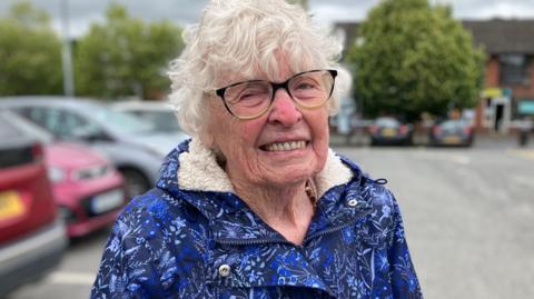 A pensioner in a blue jacket and rimmed glasses smiles for the camera