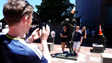 PA Media A man taking a photograph of two women and a boy wearing blue Scotland football shirts standing next to statues with orange traffic cones on their heads.