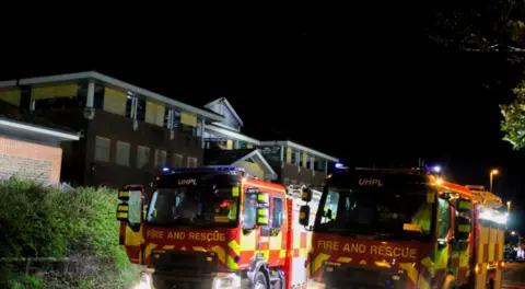 Two fire engines outside a large building with boarded up windows - it is night.