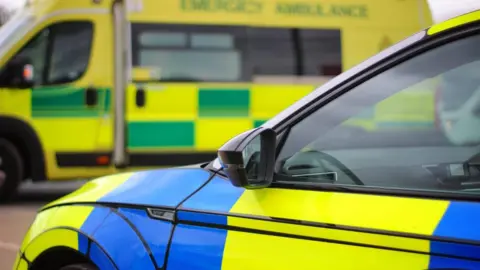 Cumbria Police Library image of a police car and ambulance. The police vehicle is in the foreground and its bonnet and right-side window can be seen. It has a yellow- and-blue colour scheme. The ambulance has a yellow-and-green livery.